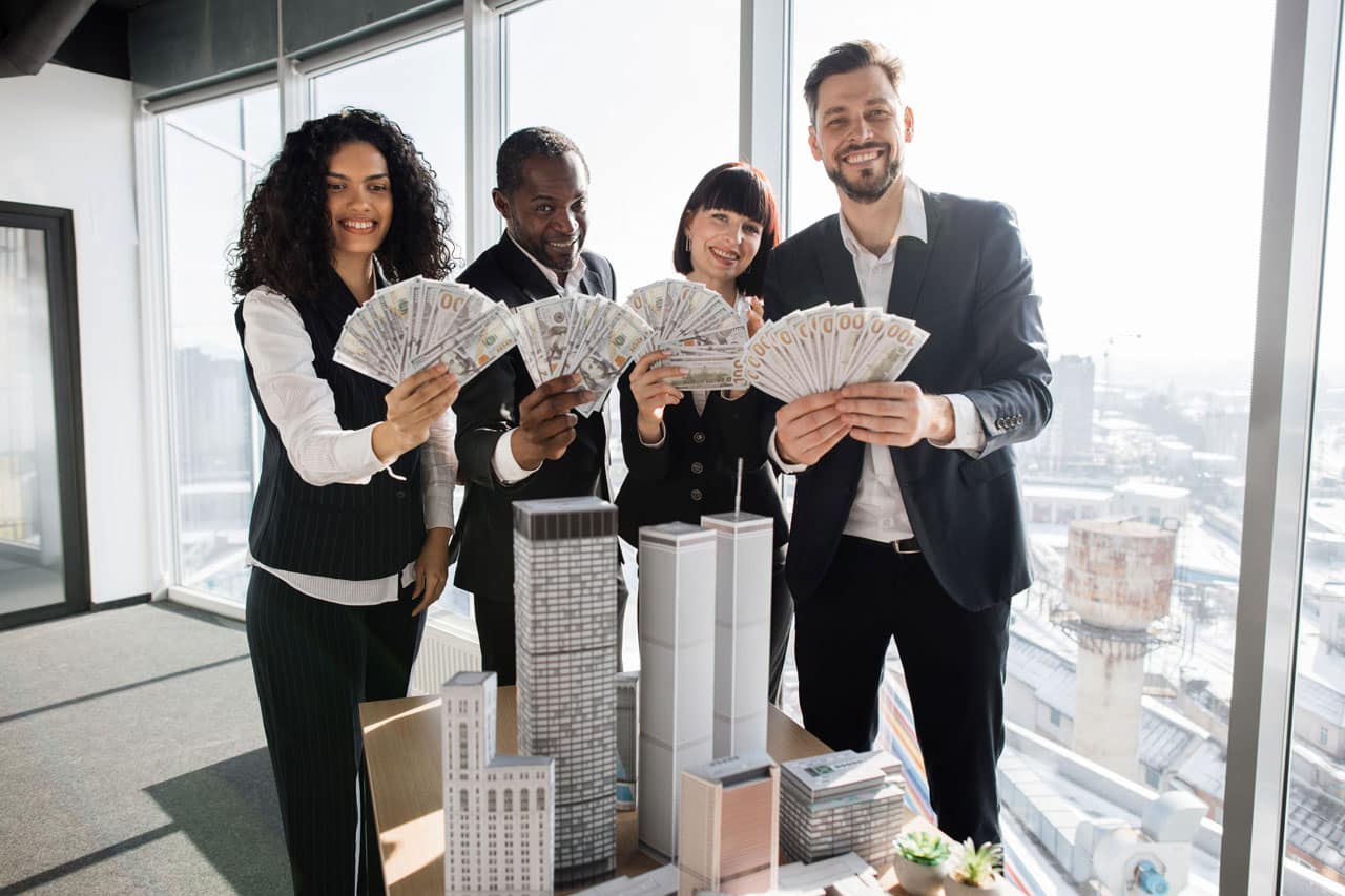Four diverse business professionals smiling and holding fanned out cash in front of miniature city buildings, symbolizing the success of building wealth.